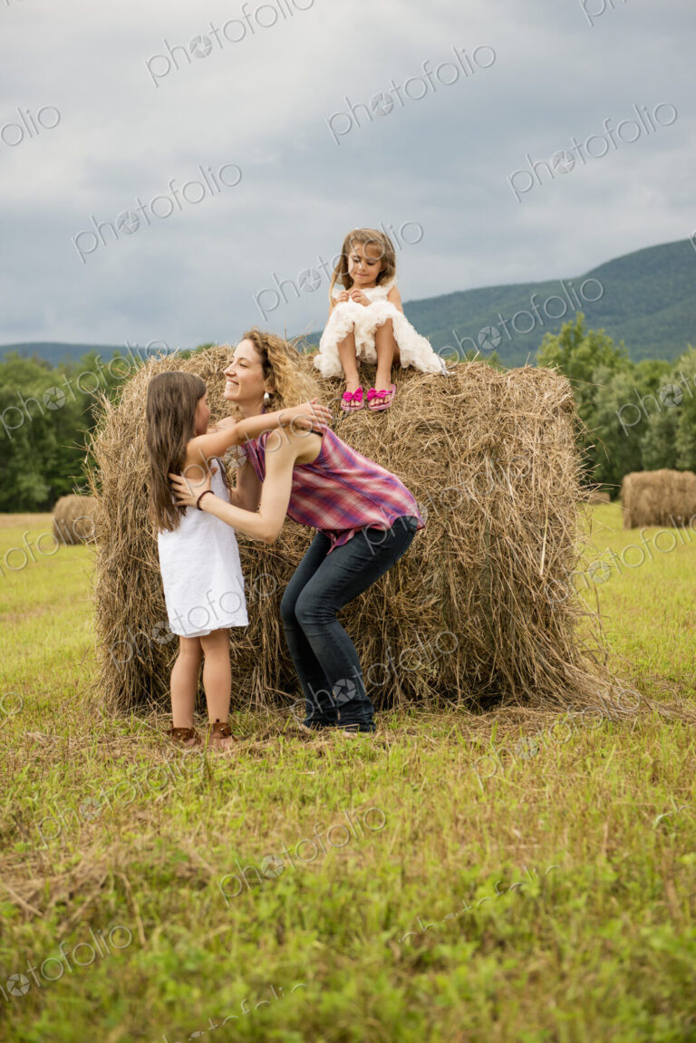 Mother playing outdoors with her daughters lifting them on top of a haystack.