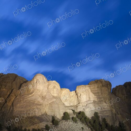 Mount Rushmore illuminated at dusk beneath a deep blue sky with soft cloud motion,…