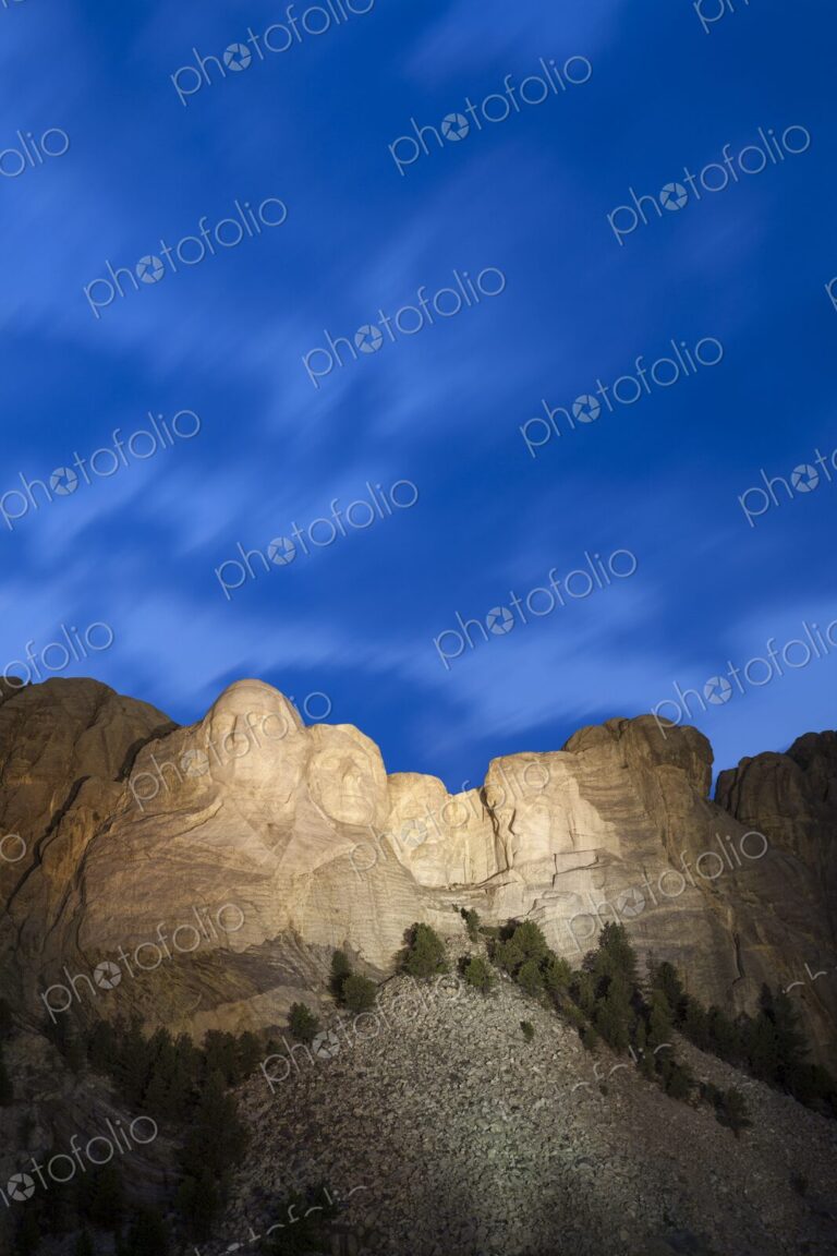 Mount Rushmore illuminated at dusk beneath a deep blue sky with soft cloud motion,…