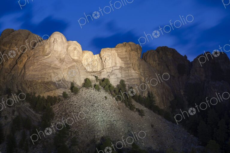 Mount Rushmore illuminated at dusk beneath a deep blue sky with soft cloud motion,…