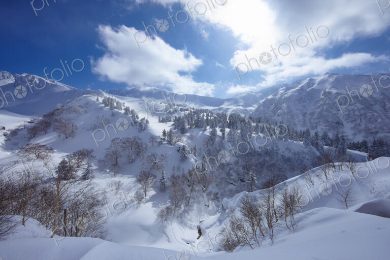 Mountainous landscape with trees on snow-covered slopes under a cloudy sky.