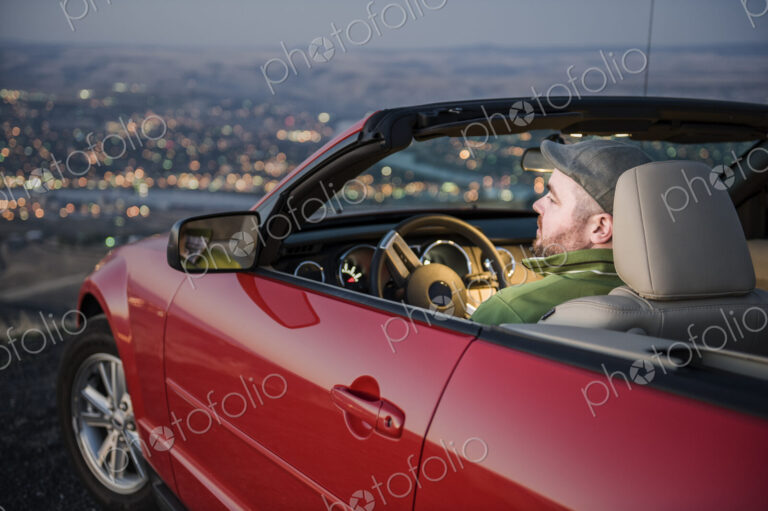 Msn parked in his convertible sports car watching the sunset over a city