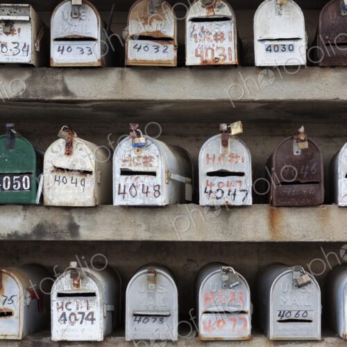 Multiple rows of weathered U.S. Mail mailboxes with hand-painted numbers are mounted… Multiple rows of weathered U.S. Mail mailboxes with hand-painted numbers are mounted…