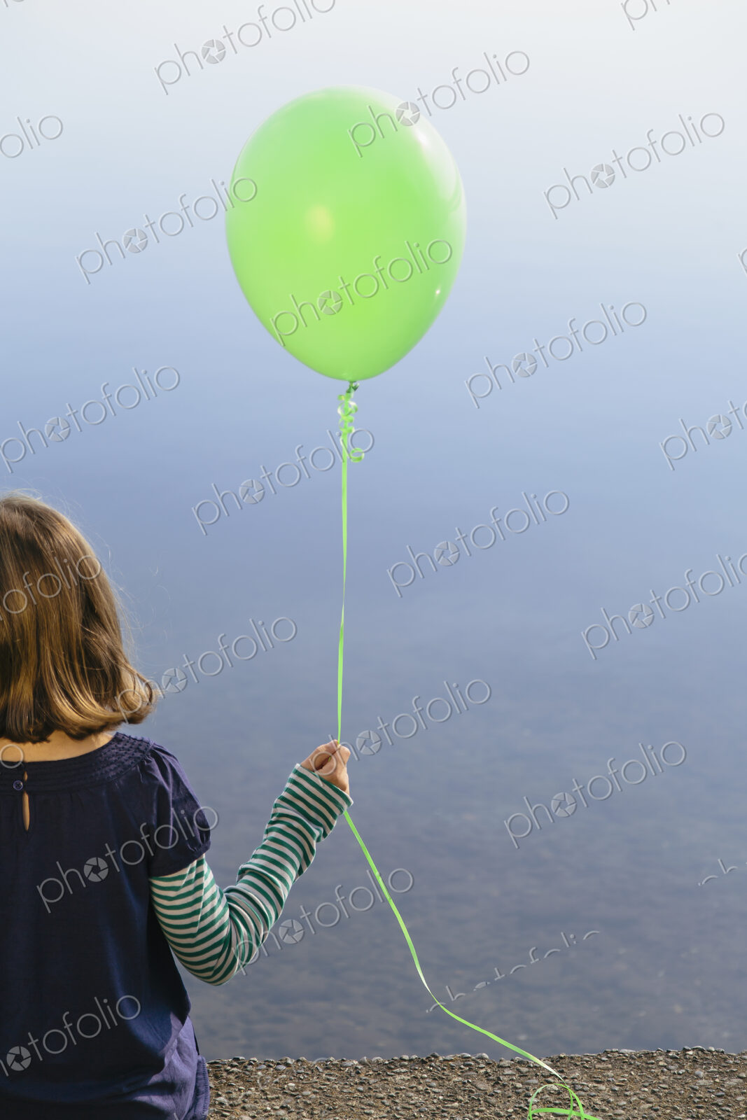 Nine year old girl holding green balloon, sitting by waters edge