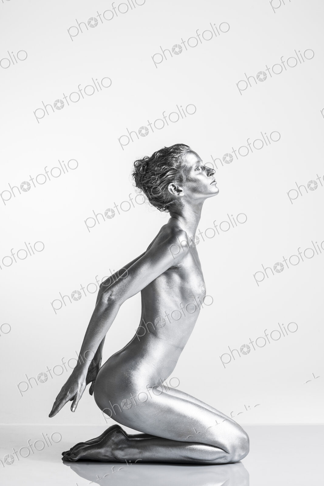 Nude Caucasian woman with silver body paint kneeling, hands behind her