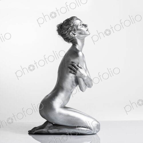 Nude Caucasian woman with silver body paint kneeling, side view, arms crossed across…