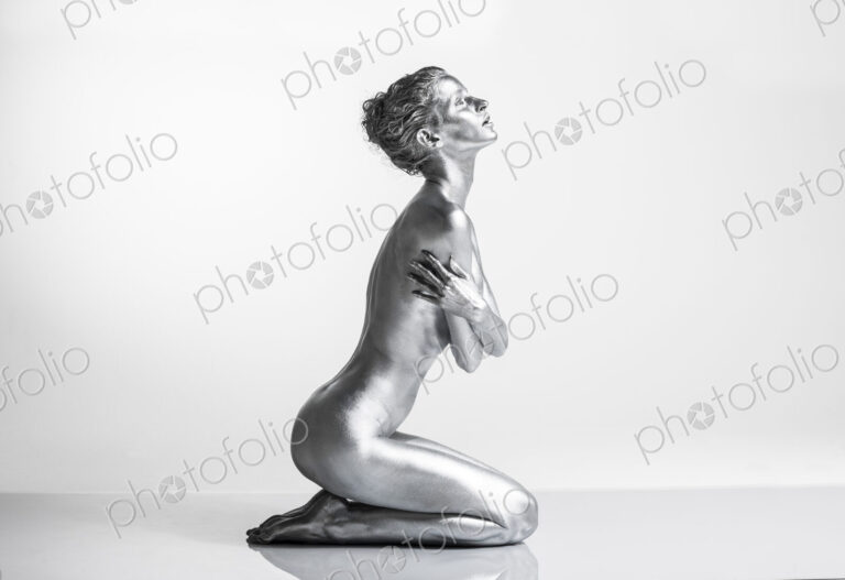 Nude Caucasian woman with silver body paint kneeling, side view, arms crossed across…