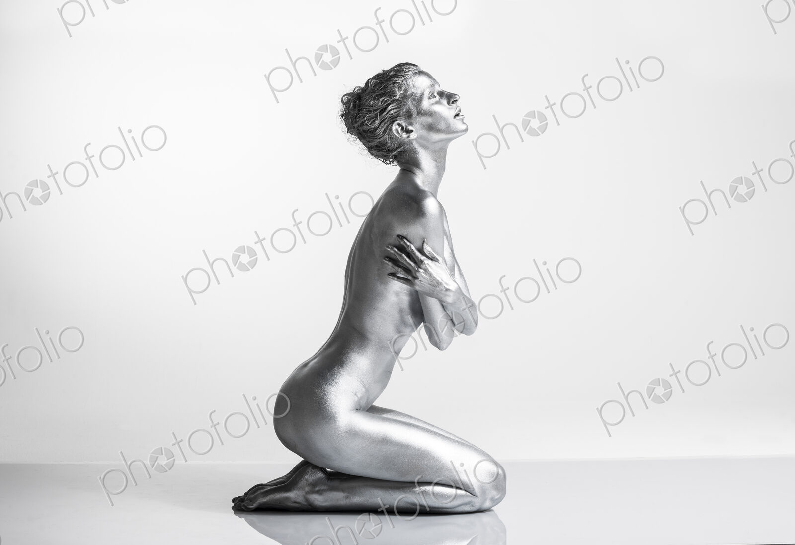Nude Caucasian woman with silver body paint kneeling, side view, arms crossed across…