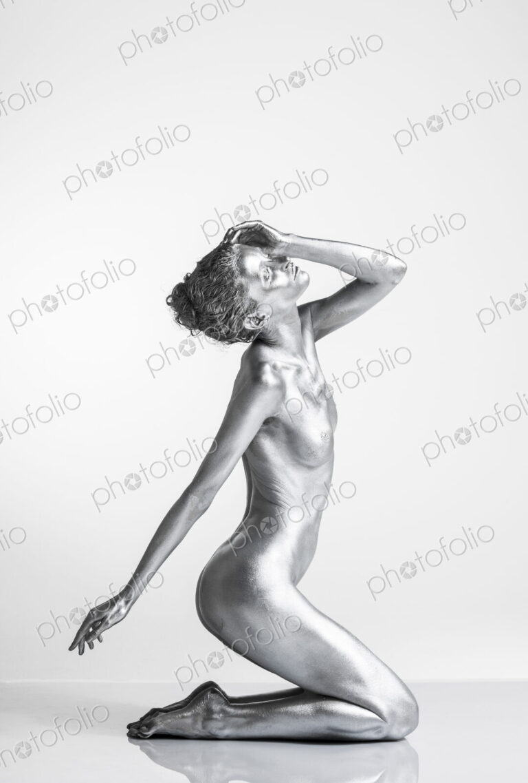 Nude Caucasian woman with silver body paint kneeling, side view hand on head