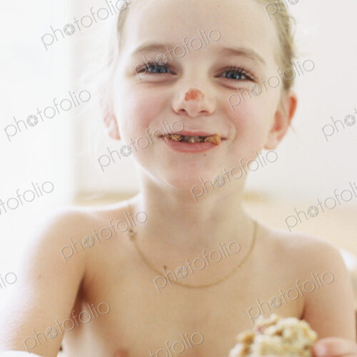 Nude young girl eating a sandwich, with crumbs in her mouth and tomato sauce on her…
