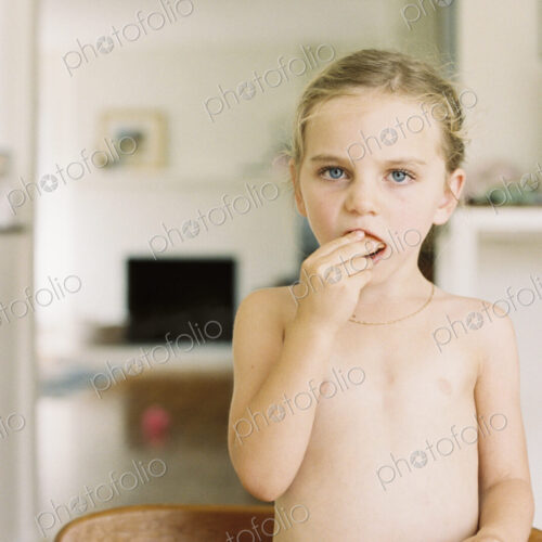 Nude young girl standing at a table, eating a sandwich and apple.
