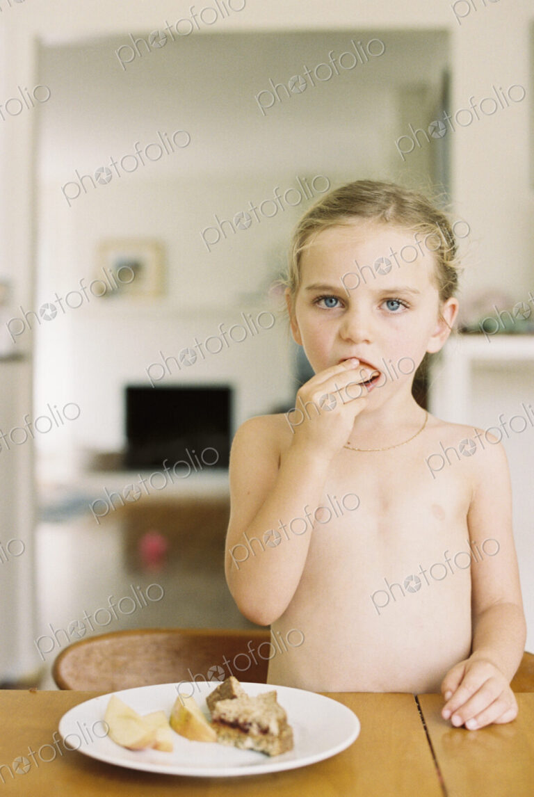Nude young girl standing at a table, eating a sandwich and apple.