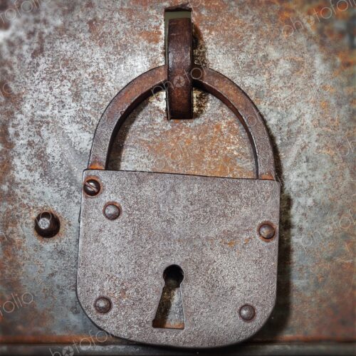 Old iron padlock hangs on a weathered metal door with rivets and rust, evoking…