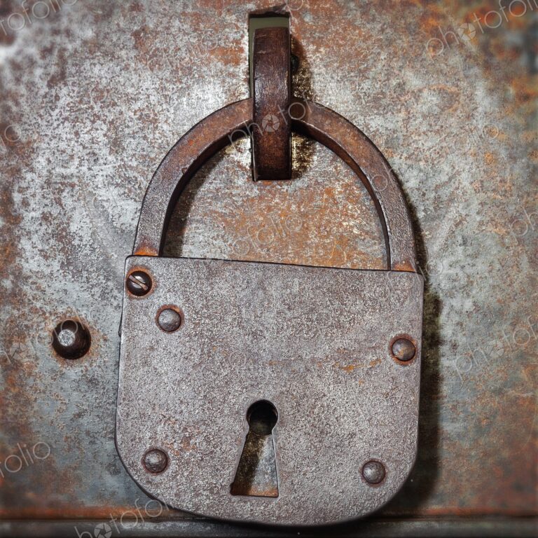 Old iron padlock hangs on a weathered metal door with rivets and rust, evoking…