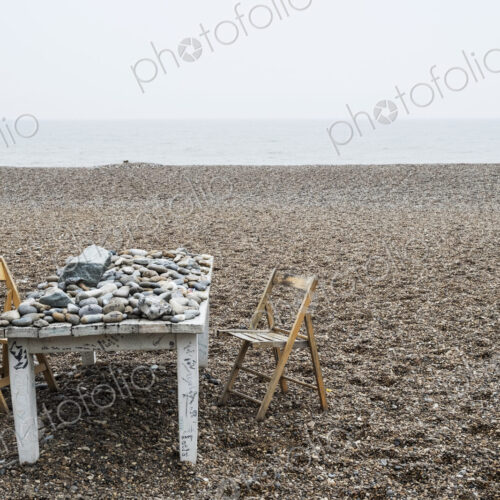 Old rickety white wooden table covered in pebbles and two folding chairs on a shingle… Old rickety white wooden table covered in pebbles and two folding chairs on a shingle…