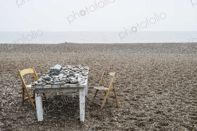 Old rickety white wooden table covered in pebbles and two folding chairs on a shingle…