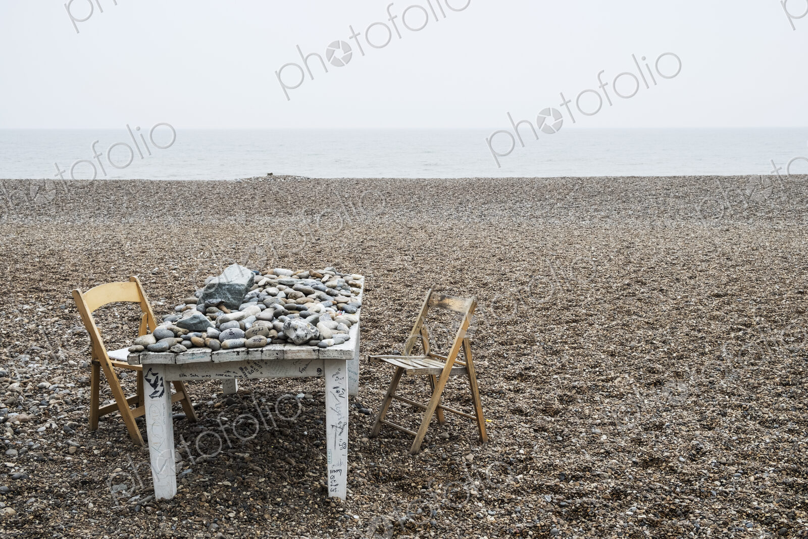 Old rickety white wooden table covered in pebbles and two folding chairs on a shingle…