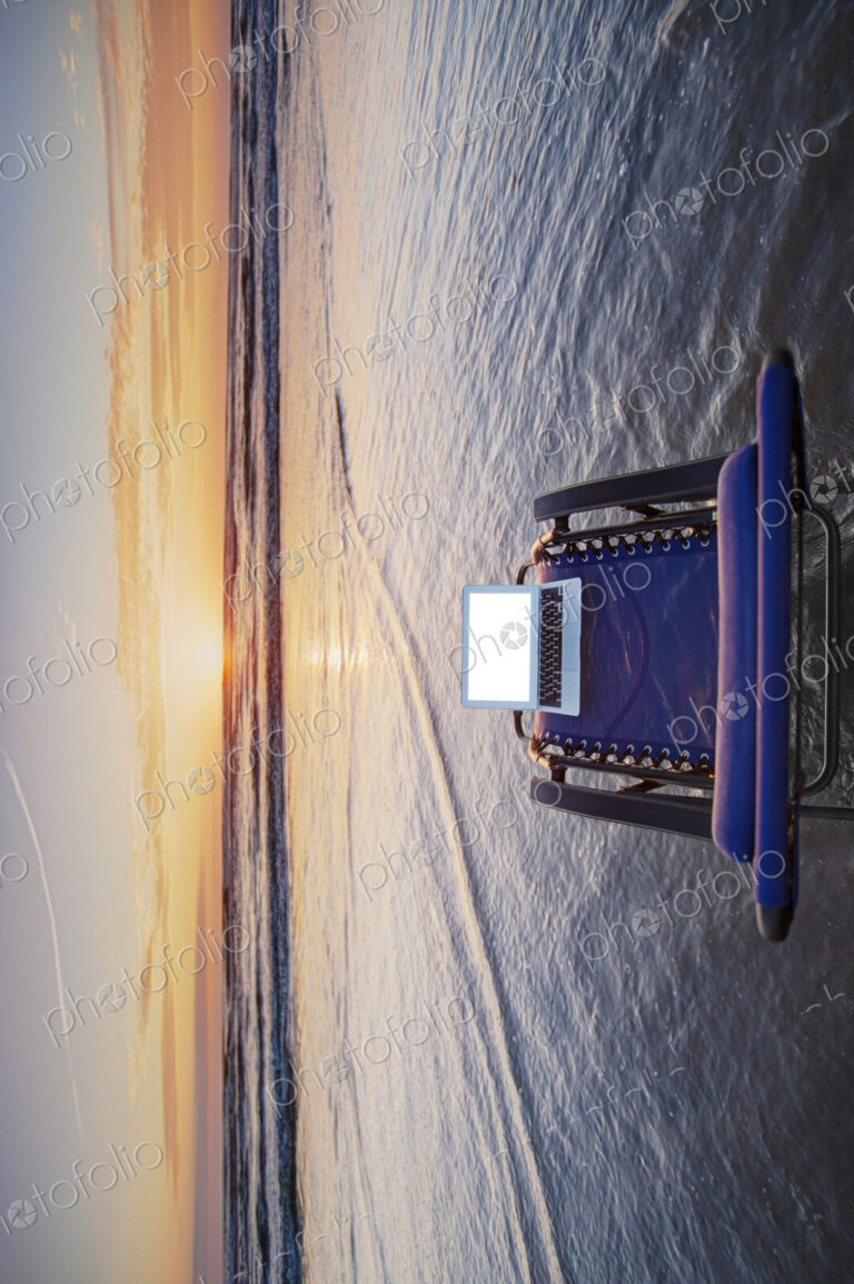 Open laptop on sun lounger in shallow wave at beach at sunset.