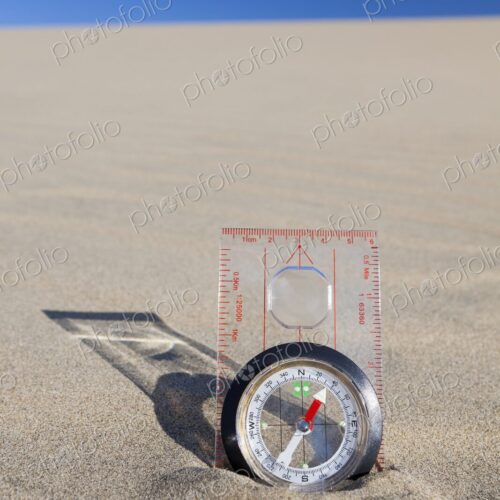 Orienteering compass placed upright in a sand dune beneath a clear blue sky,…