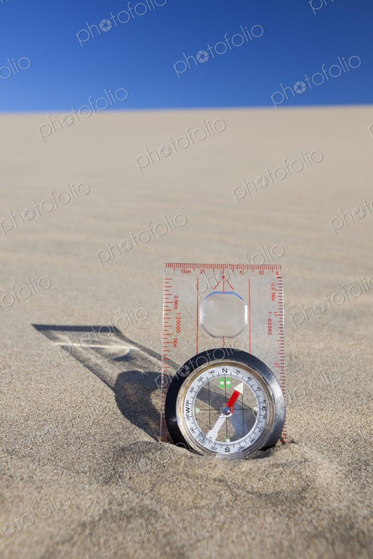 Orienteering compass placed upright in a sand dune beneath a clear blue sky,…
