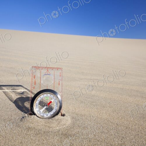 Orienteering compass placed upright in a sand dune beneath a clear blue sky,…