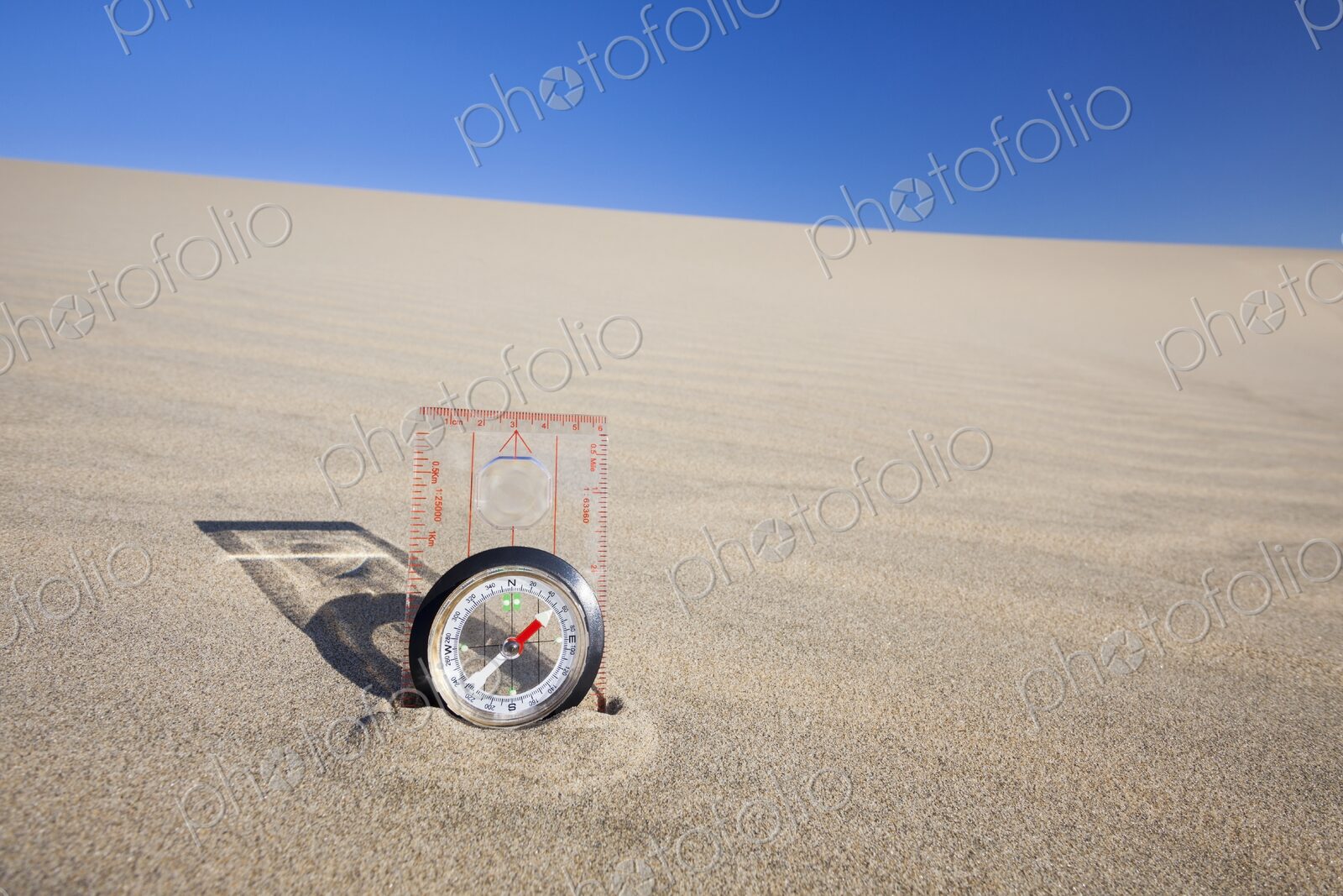 Orienteering compass placed upright in a sand dune beneath a clear blue sky,…