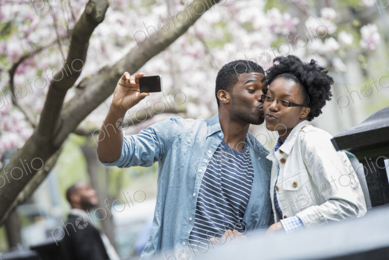 Outdoors in the city in spring. An urban lifestyle. A man kissing a woman and taking a…