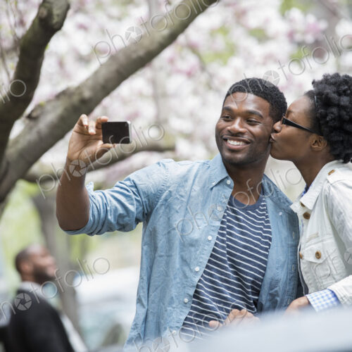 Outdoors in the city in spring. An urban lifestyle. A woman kissing a man and taking a…