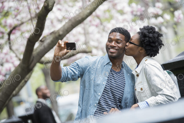 Outdoors in the city in spring. An urban lifestyle. A woman kissing a man and taking a…