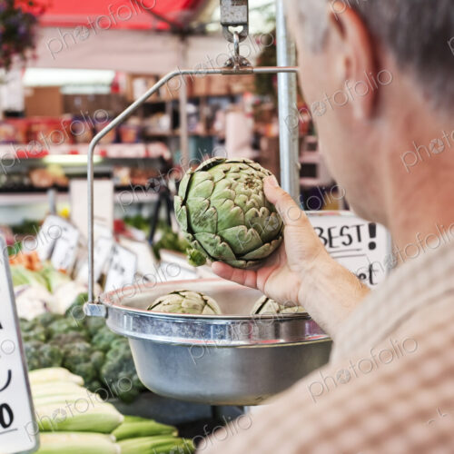 Over the shoulder view of man weighing artichoke at a fruit and vegetable market. Over the shoulder view of man weighing artichoke at a fruit and vegetable market.