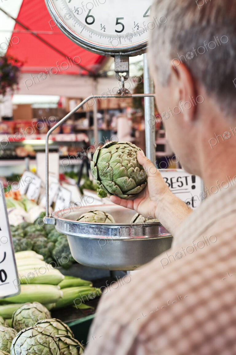 Over the shoulder view of man weighing artichoke at a fruit and vegetable market.
