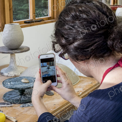 Over the shoulder view of woman sitting in her ceramics workshop, checking her mobile…