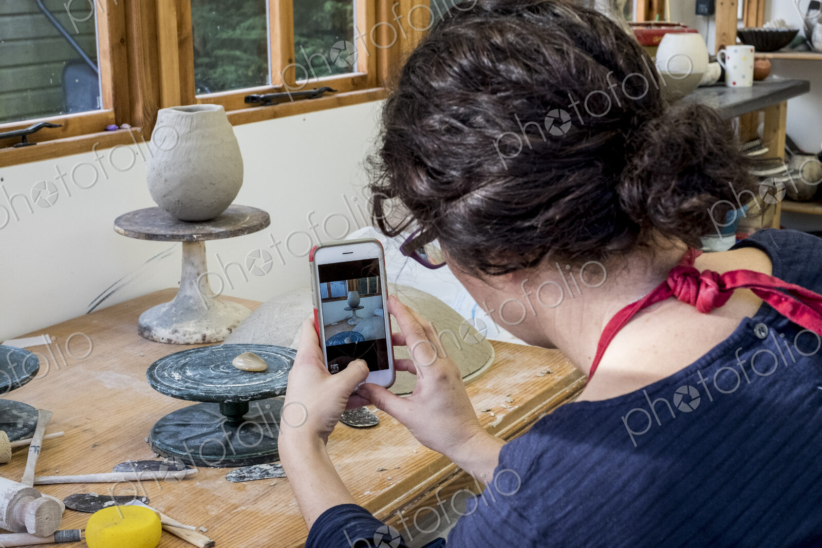 Over the shoulder view of woman sitting in her ceramics workshop, checking her mobile…