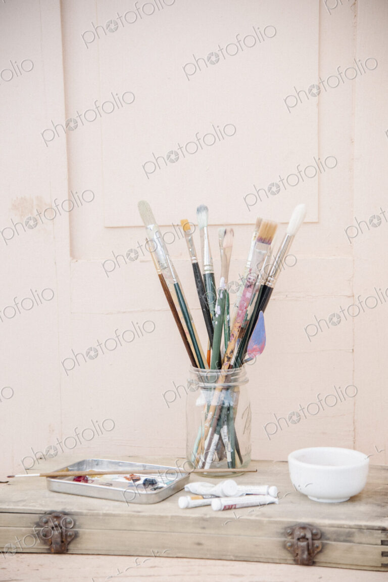 Painting materials, glass jar with paint brushes, paint tubes and a bowl.