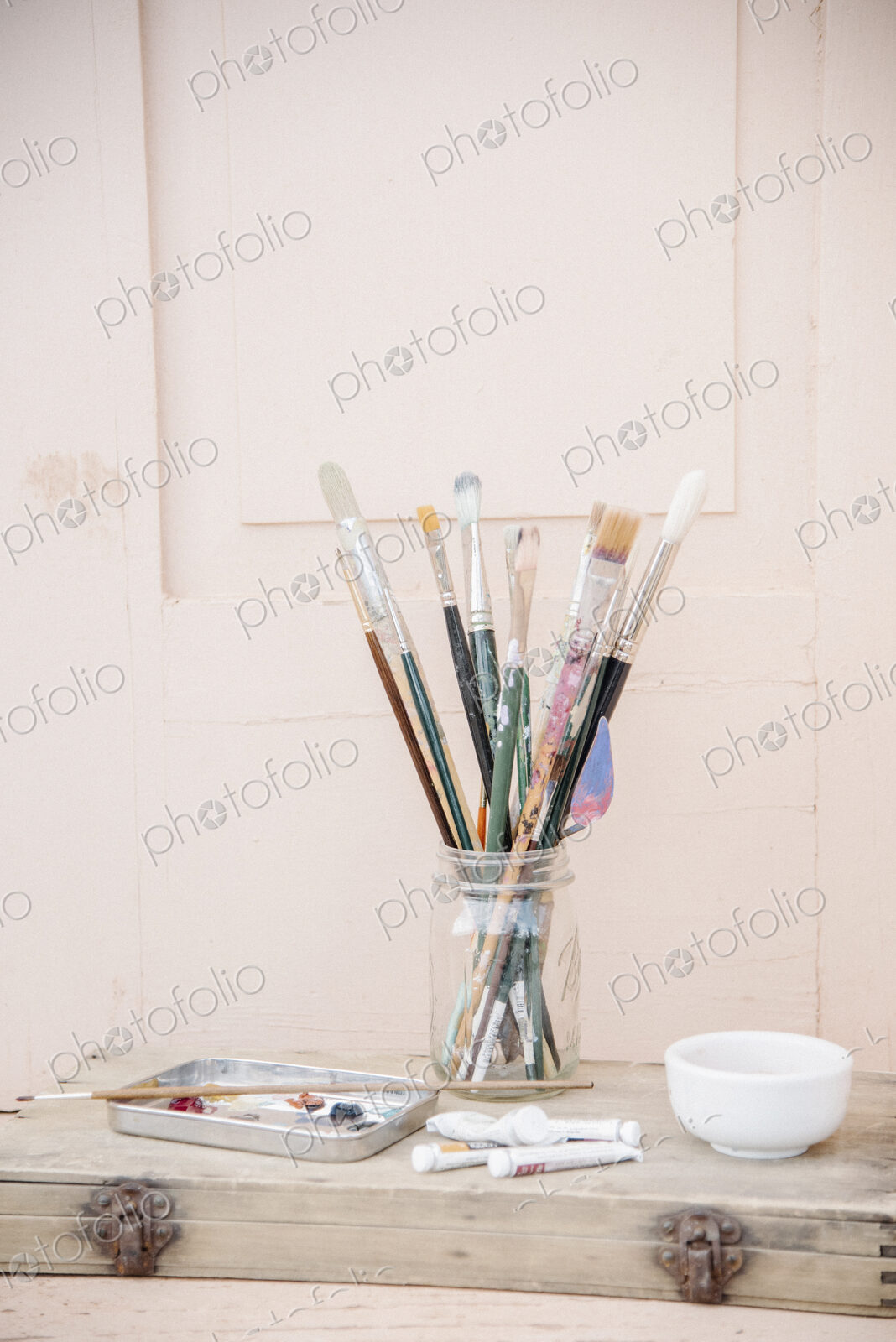 Painting materials, glass jar with paint brushes, paint tubes and a bowl.