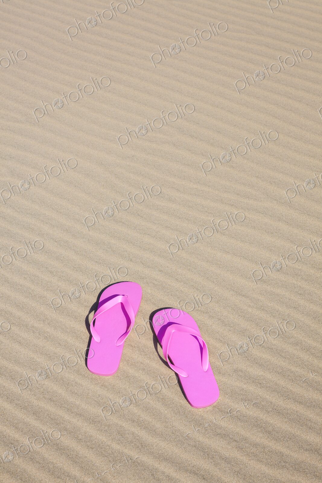 Pair of bright pink flip flops resting on smooth, rippled sand under the sun.