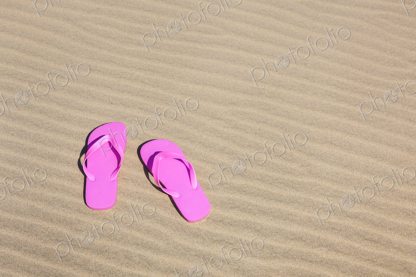 Pair of bright pink flip flops resting on smooth, rippled sand under the sun.