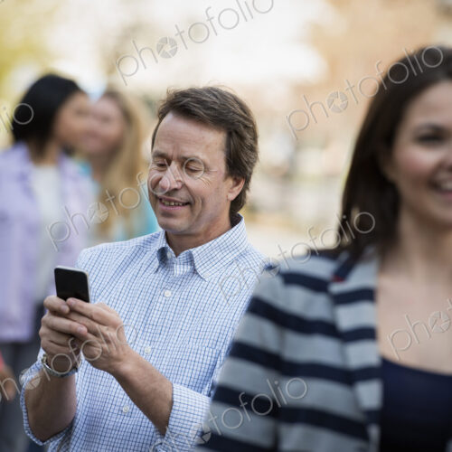 People outdoors in the city in spring time. A man checking his cell phone, among a… People outdoors in the city in spring time. A man checking his cell phone, among a…