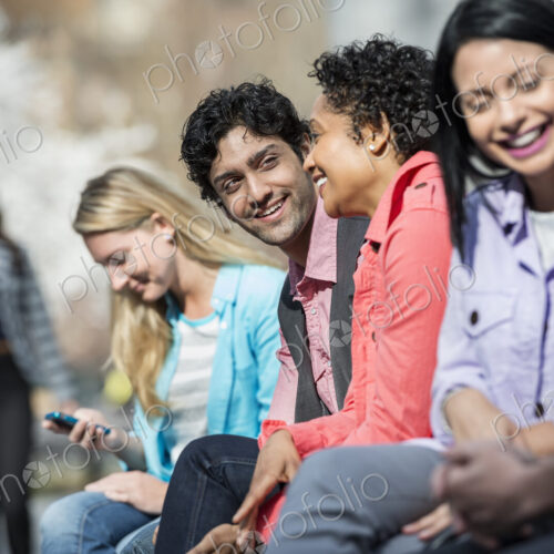 People outdoors in the city in spring time. Five people sitting in a row, one looking… People outdoors in the city in spring time. Five people sitting in a row, one looking…