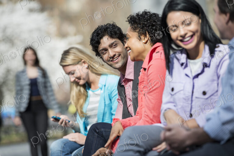 People outdoors in the city in spring time. Five people sitting in a row, one looking…