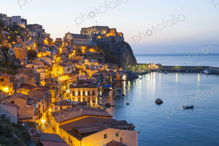 Picturesque coastal town at dusk, with illuminated castle on cliff.