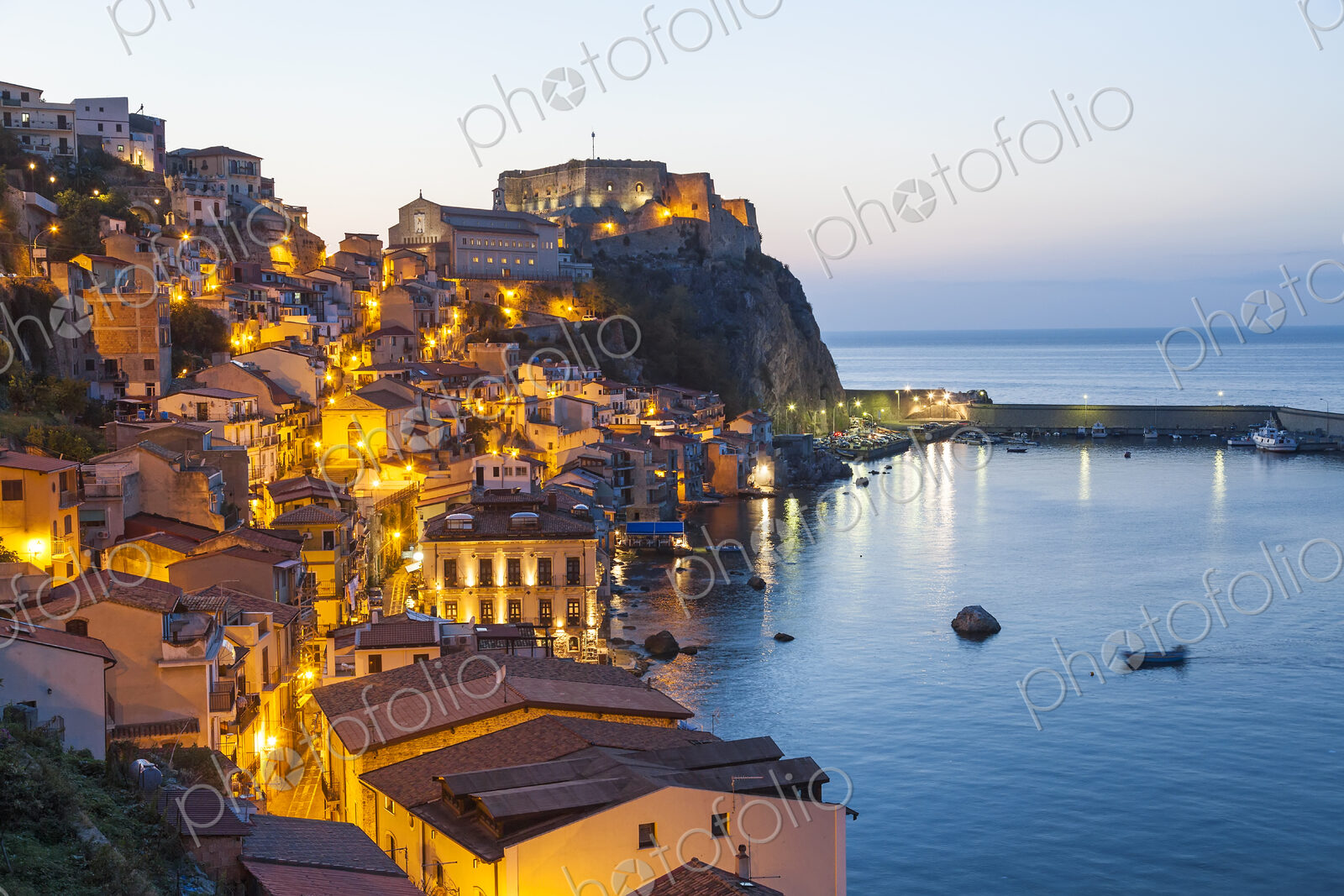 Picturesque coastal town at dusk, with illuminated castle on cliff.