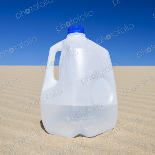 Plastic gallon water jug resting on sunlit sand dune with a vibrant blue sky overhead.…