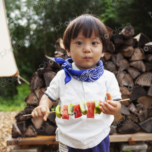 Portrait of Japanese boy holding skewer with fresh kiwi and melon, looking at camera.