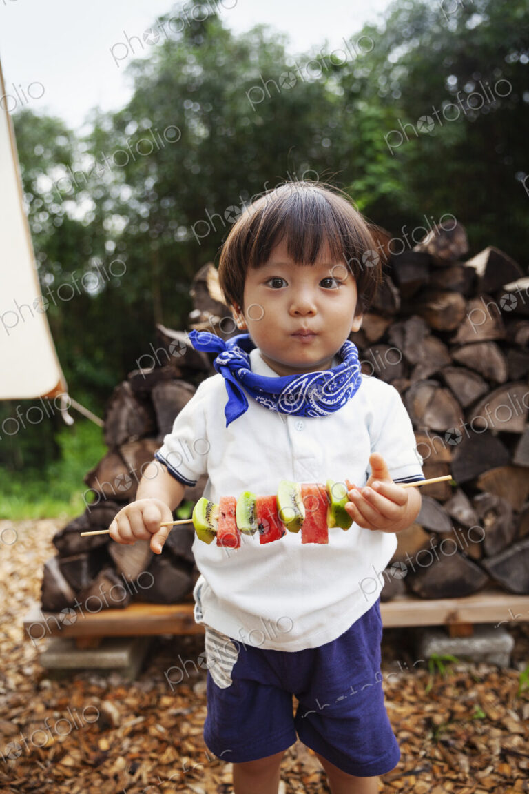 Portrait of Japanese boy holding skewer with fresh kiwi and melon, looking at camera.