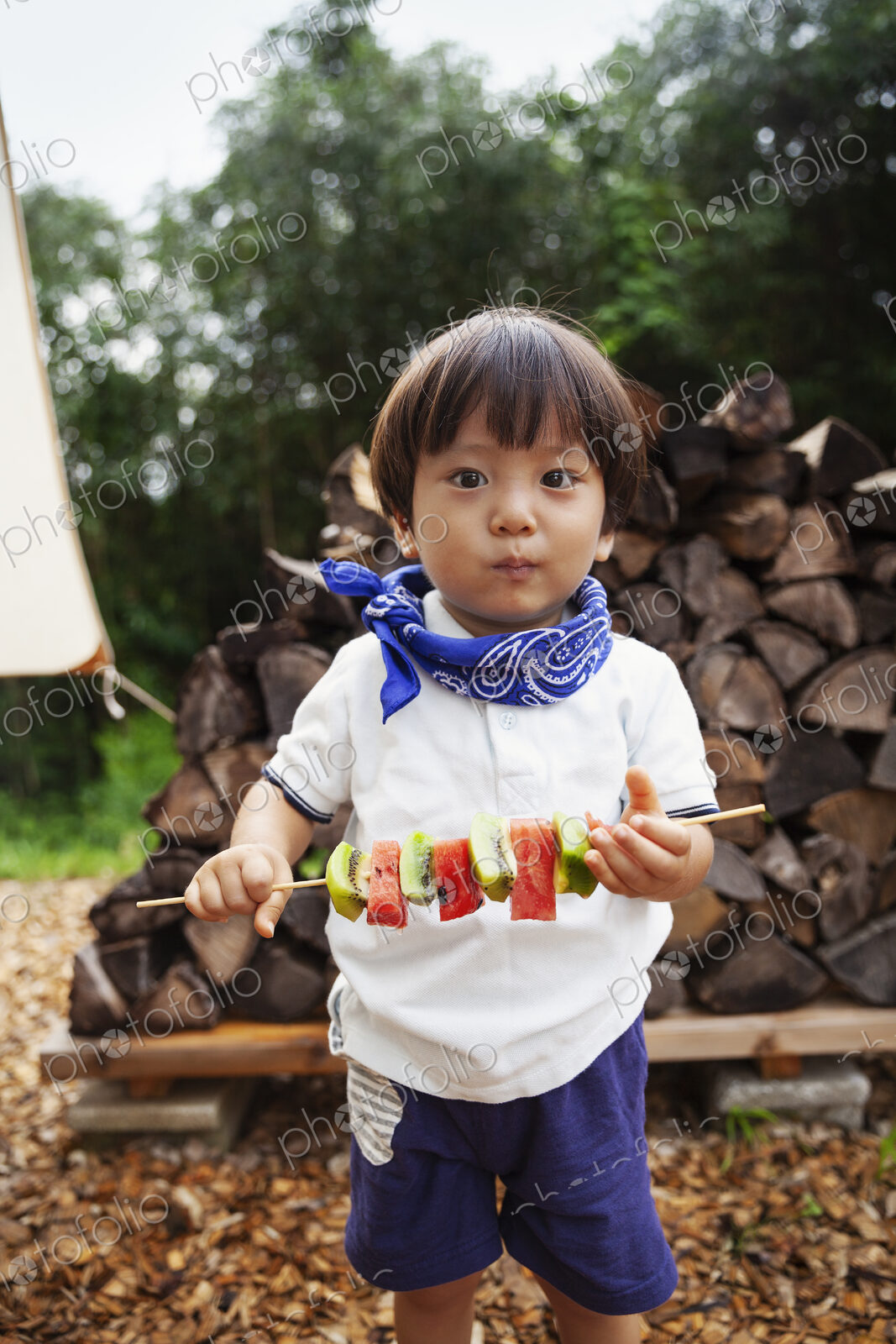 Portrait of Japanese boy holding skewer with fresh kiwi and melon, looking at camera.