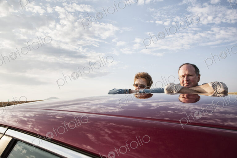 Portrait of a Caucasian senior couple leaning on their car at a rest stop on a road trip.