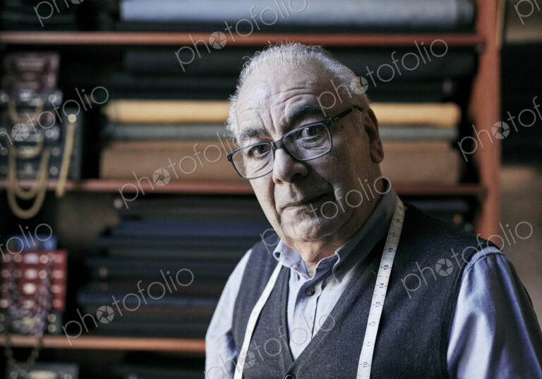 Portrait of a senior tailor with a measuring tape around his neck, in his workroom.