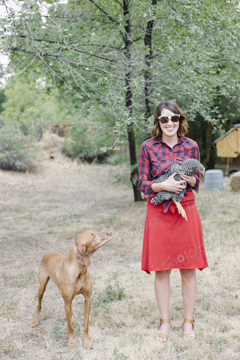 Portrait of a smiling woman holding a grey hen, a dog standing beside her.