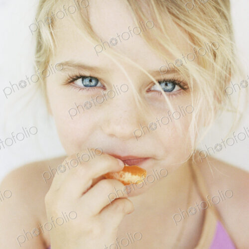 Portrait of a young blond girl, eating a mandarin, looking at the camera.